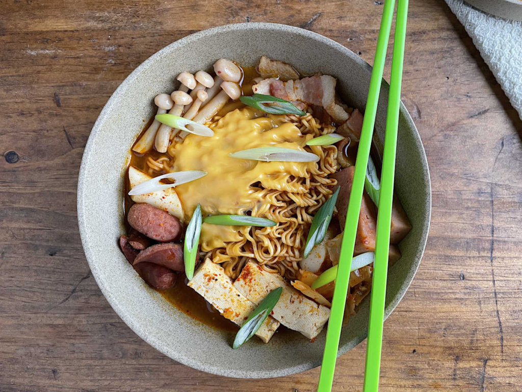 Korean Army Stew in a tan bowl with green chopsticks on top