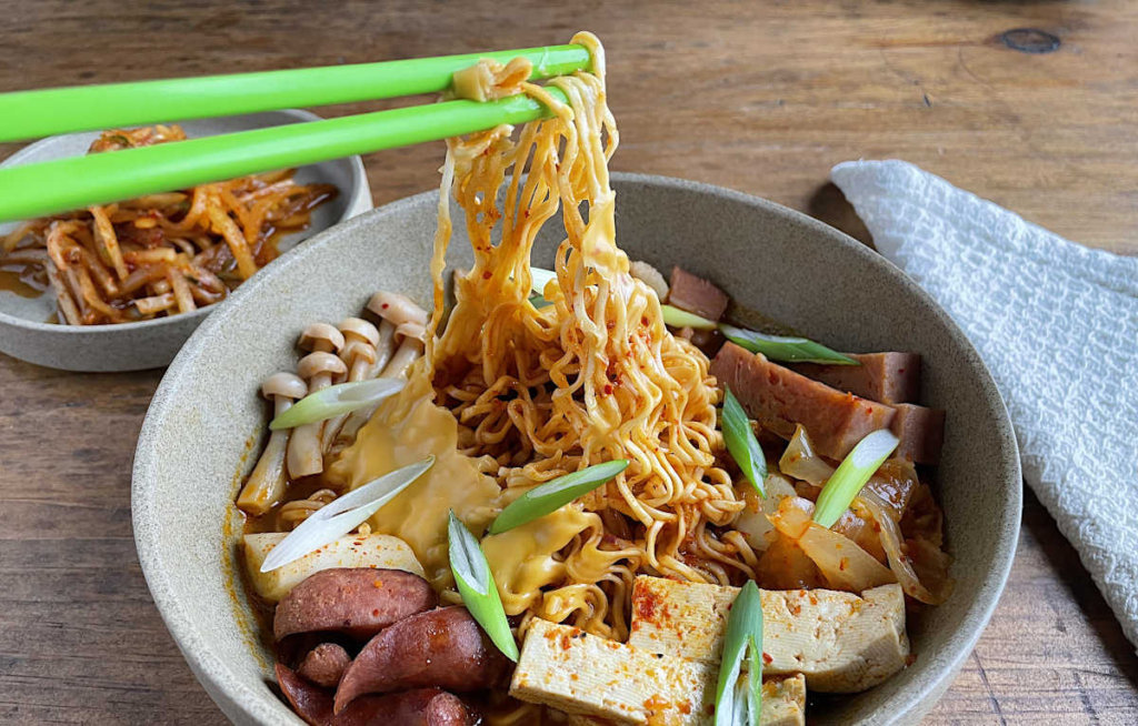 Korean Army Stew with noodles held in green chopsticks